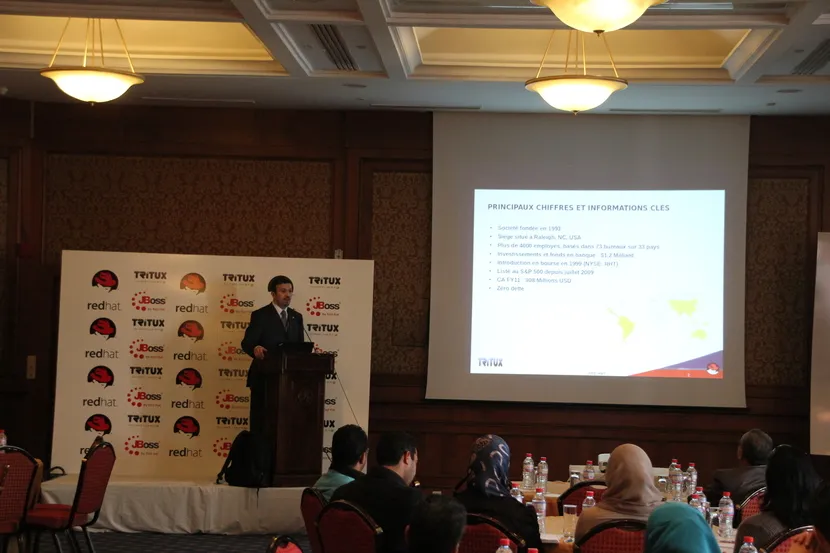 A speaker at a lectern in front of a Red Hat, JBoss and Tritux backdrop, presenting a slide of Red Hat's key figures to the seminar audience
