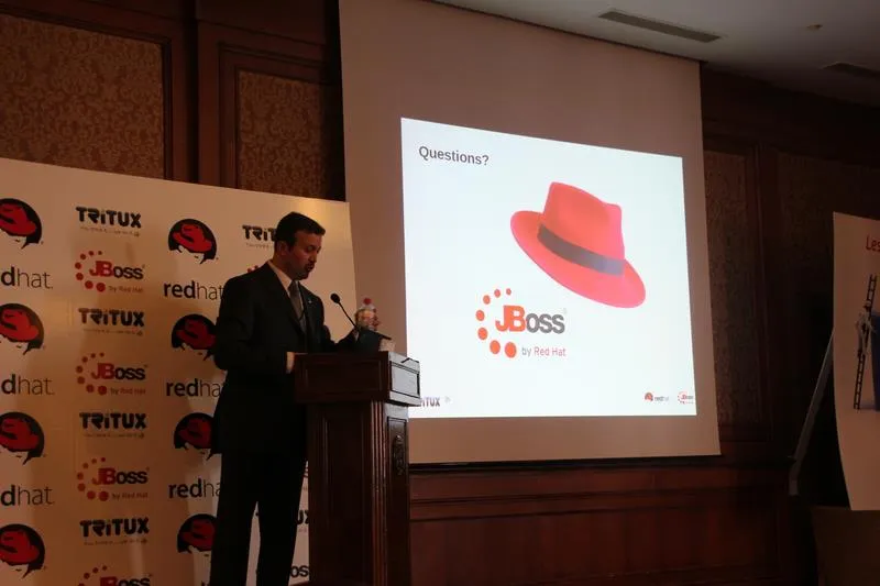 A speaker at the lectern in front of a closing slide reading "Questions?" with the Red Hat fedora and the JBoss by Red Hat logo